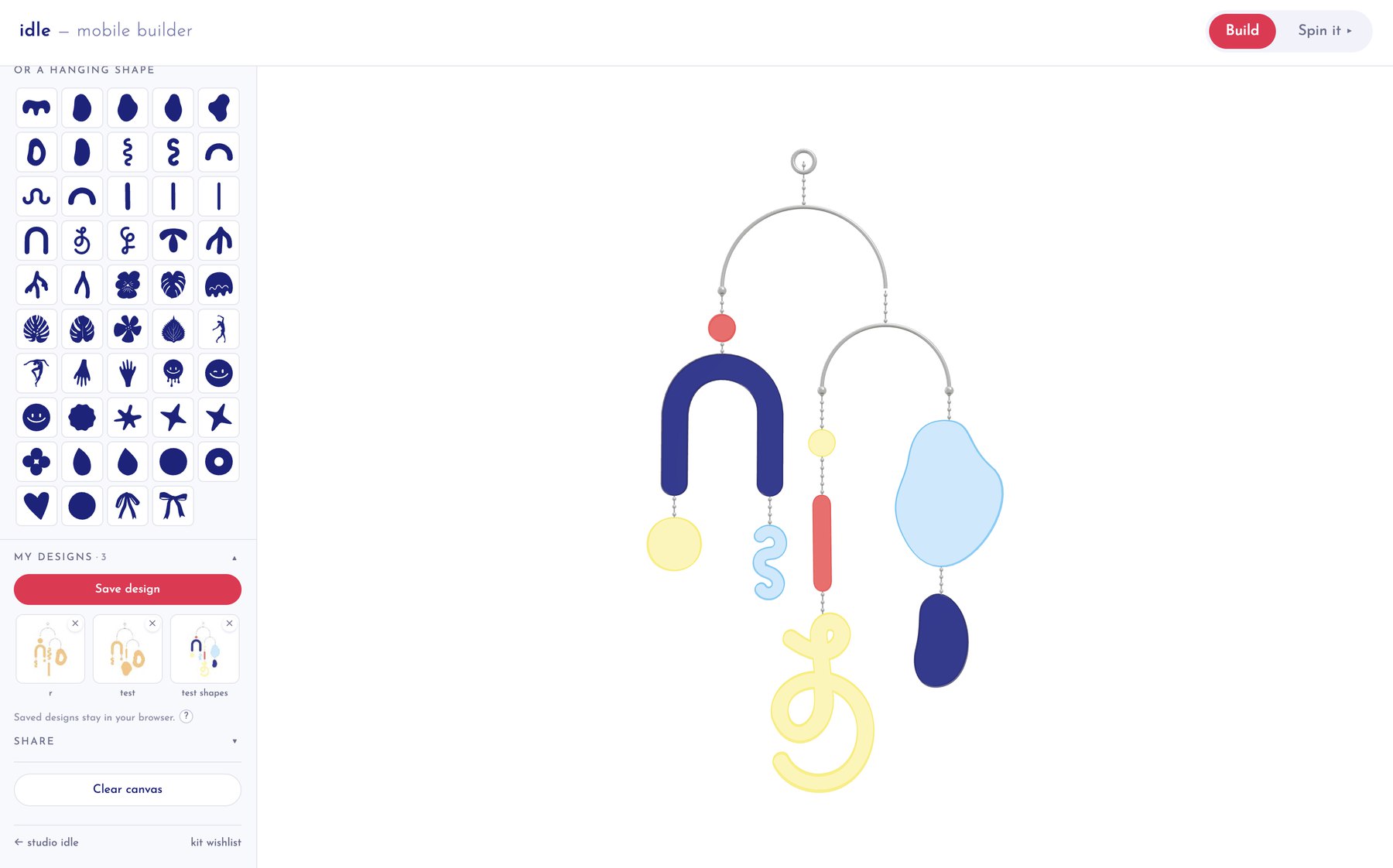 The Idle Mobile Builder in a browser: a scrolling library of navy shapes down the left with a saved-designs panel beneath it, Build and Spin it controls in the header, and a two-tier mobile hanging in the canvas with navy, yellow, red and pale blue shapes on beaded chains